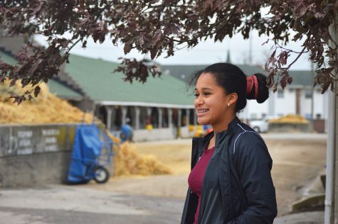 A photo of a young woman smiling in front of the stables inside of Churchill Downs. The young woman is part of the Backside Learning Center.